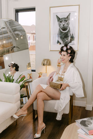 Woman in white satin robe with hair rollers and pearl necklace sits in a plush armchair in a chic salon-style dressing room, holding a vintage gold telephone beneath a framed cat portrait and a large mirror reflection.