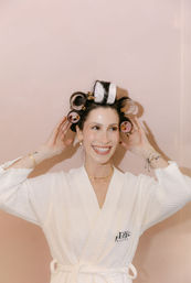 Smiling woman in a white spa robe with pink and white hair rollers, adjusting them and showing gold jewelry against a pale pink background — playful beauty routine portrait.