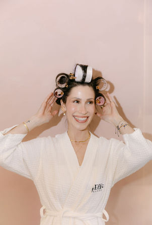 Smiling woman in a white spa robe with pink and white hair rollers, adjusting them and showing gold jewelry against a pale pink background — playful beauty routine portrait.