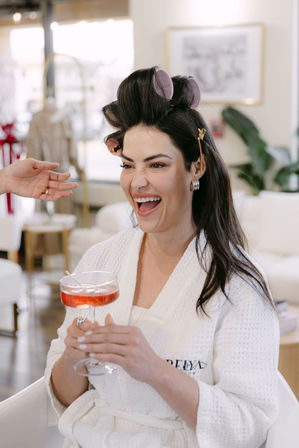 Laughing woman in a white robe with hair rollers and clips, holding a pink cocktail while getting styled in a bright salon