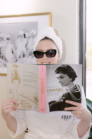 Person in a white bathrobe and towel turban wearing sunglasses, smiling while reading a vintage fashion book with a black-and-white portrait, seated in a bright spa-like room