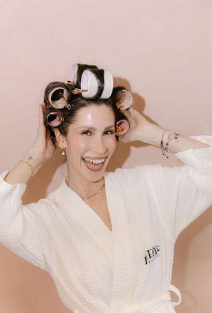 Smiling woman in a white spa robe wearing pink and white hair rollers, adjusting curls against a pastel pink background — playful beauty routine.