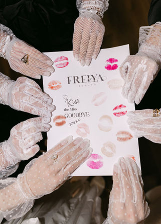Overhead shot of hands in white lace gloves holding a keepsake card stamped with colorful lipstick kiss marks and the phrase “Kiss the Miss Goodbye,” playful bridal party bachelorette image.