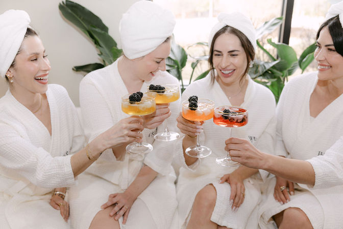 Four friends in white spa robes and towel turbans clinking fruity cocktails topped with blackberries in a bright indoor spa lounge with plants.