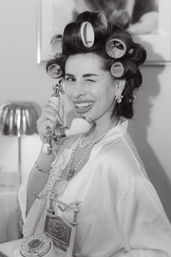 Black-and-white retro photo of a woman in hair curlers and pearls, winking while holding a vintage rotary telephone handset in a glamorous robe.