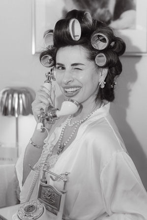 Black-and-white retro photo of a woman in hair curlers and pearls, winking while holding a vintage rotary telephone handset in a glamorous robe.