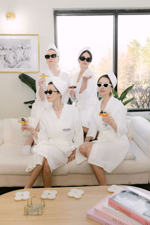 Four women in white spa robes and towel turbans wearing sunglasses, lounging on a bright living-room sofa and sipping cocktails during a playful at-home spa day.