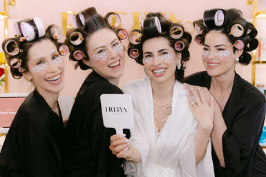Four women in a beauty salon wearing hair rollers and under-eye patches, laughing in black and white robes during a playful getting-ready beauty session as one holds a handheld mirror.