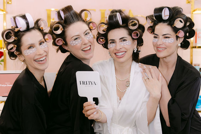 Four women in a beauty salon wearing hair rollers and under-eye patches, laughing in black and white robes during a playful getting-ready beauty session as one holds a handheld mirror.