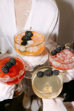 Four elegant coupe cocktails with ice and blackberry skewers in red, orange and pale-yellow hues, held by gloved hands at a chic cocktail party or brunch.