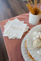 Stack of white cocktail napkins with gold foil lettering on a dusty pink tablecloth, beside a textured white cup of wooden stirrers and a tiered white dessert plate — elegant event display