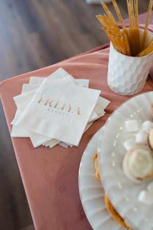 Stack of white cocktail napkins with gold foil lettering on a dusty pink tablecloth, beside a textured white cup of wooden stirrers and a tiered white dessert plate — elegant event display