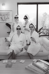 Black-and-white image of four women in white bathrobes and towel turbans wearing sunglasses, lounging on a sofa and toasting with champagne in a chic at-home spa lounge by a large window with plants and a coffee table stacked with fashion books.