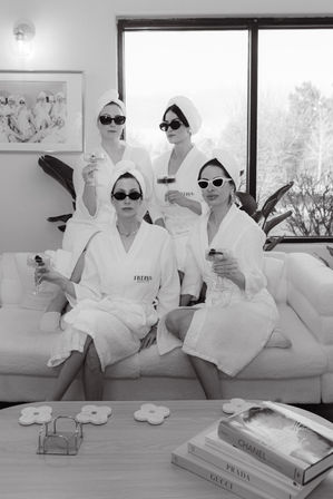Black-and-white image of four women in white bathrobes and towel turbans wearing sunglasses, lounging on a sofa and toasting with champagne in a chic at-home spa lounge by a large window with plants and a coffee table stacked with fashion books.