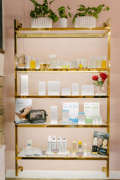 Gold-framed retail shelf against a soft pink wall showcasing skincare bottles, boxed products, and jars with potted green plants on top and two red roses — bright spa-style display.