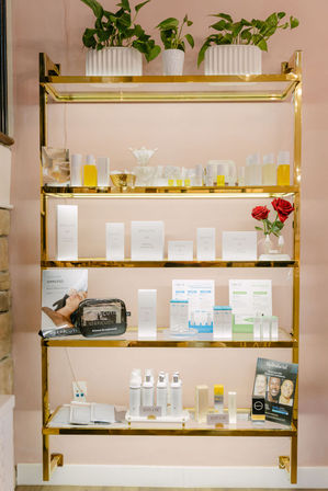 Gold-framed retail shelf against a soft pink wall showcasing skincare bottles, boxed products, and jars with potted green plants on top and two red roses — bright spa-style display.