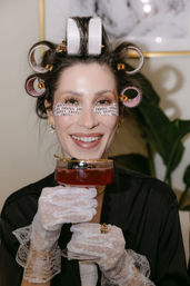 Smiling woman in hair rollers and printed under-eye patches wearing white lace gloves and a black robe, holding a red cocktail in a coupe glass during an at-home spa night