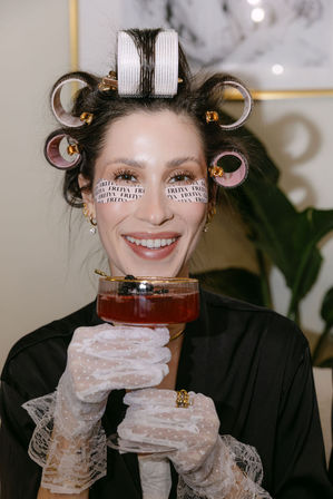Smiling woman in hair rollers and printed under-eye patches wearing white lace gloves and a black robe, holding a red cocktail in a coupe glass during an at-home spa night