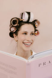 Smiling woman with pink and white hair rollers reading a pink fashion book against a soft blush background — playful at-home beauty routine