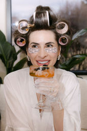 Woman in a white robe with pink hair rollers and printed under-eye patches sipping a peach cocktail from a coupe, wearing sheer polka-dot gloves and pearl jewelry during a playful at-home beauty prep.