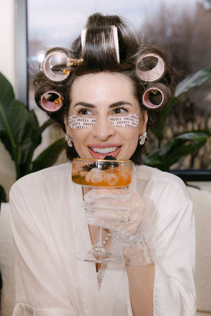 Woman in a white robe with pink hair rollers and printed under-eye patches sipping a peach cocktail from a coupe, wearing sheer polka-dot gloves and pearl jewelry during a playful at-home beauty prep.