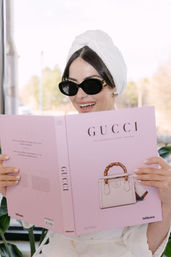 Smiling person in a white towel turban and black sunglasses reading a pink luxury-fashion coffee table book with a handbag on the cover in a bright indoor setting.