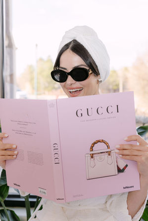Smiling person in a white towel turban and black sunglasses reading a pink luxury-fashion coffee table book with a handbag on the cover in a bright indoor setting.