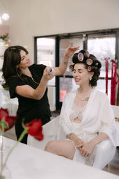Bridal client in a white robe with pearl necklace sits with pink hair rollers while a stylist sprays hairspray in a bright beauty salon during wedding morning prep.