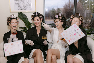 Bridal pamper party: four women in satin robes and hair rollers laughing on a sofa during getting-ready spa time, wearing under-eye patches and lace gloves, holding champagne flutes and bridal-themed gift boxes in a bright living room with plants and a large window.