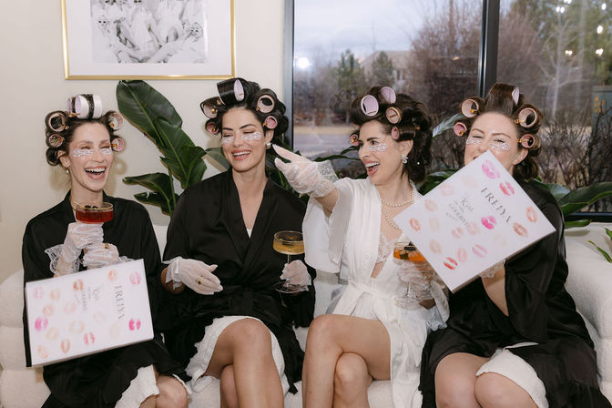Bridal pamper party: four women in satin robes and hair rollers laughing on a sofa during getting-ready spa time, wearing under-eye patches and lace gloves, holding champagne flutes and bridal-themed gift boxes in a bright living room with plants and a large window.