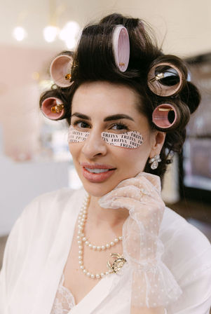 Smiling woman in a beauty salon with pink hair rollers and under-eye patches, wearing pearls and lace glove during bridal glam prep