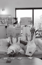 Black-and-white photo of three people in white spa robes lounging on a couch, faces hidden behind oversized fashion coffee-table books, with cocktail glasses on a coffee table and a windowed living-room backdrop.