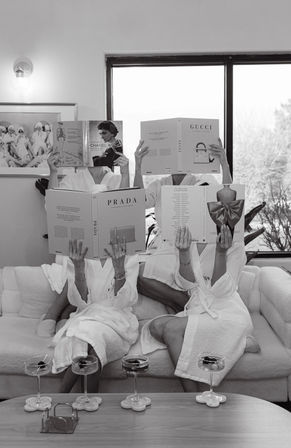 Black-and-white photo of three people in white spa robes lounging on a couch, faces hidden behind oversized fashion coffee-table books, with cocktail glasses on a coffee table and a windowed living-room backdrop.