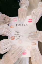 Top-down view of several hands in white lace and mesh gloves holding a card stamped with colorful lipstick kiss marks and the words Kiss the Miss Goodbye — playful bachelorette/bridal shower moment.