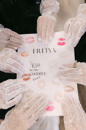 Top-down view of several hands in white lace and mesh gloves holding a card stamped with colorful lipstick kiss marks and the words Kiss the Miss Goodbye — playful bachelorette/bridal shower moment.