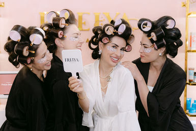 Four women in robes wearing large hair rollers and under-eye patches laugh and pose in a salon while one holds a handheld mirror during a playful beauty get-ready.