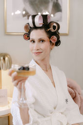 Smiling woman in a white waffle robe with pink hair rollers toasting with a champagne coupe in a bright spa-like vanity setting.
