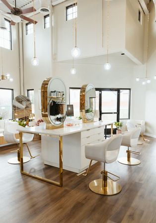 Bright modern hair salon interior with high ceilings, white-and-gold styling island, round gold-framed mirrors, white swivel styling chairs, hanging globe pendant lights, wood flooring and large windows