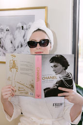 Woman in a white spa robe and towel turban wearing black cat-eye sunglasses, reading a fashion biography with a vintage black-and-white portrait on the cover in a bright spa-like room.