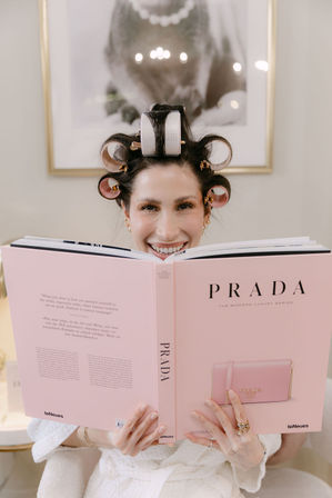 Smiling woman with hair rollers peeking over a pink luxury fashion coffee-table book while relaxing in a bright beauty salon