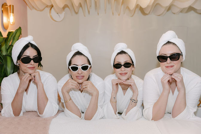 Four women in white spa robes and towel turbans wearing sunglasses, leaning on their elbows and smiling on a treatment table in a bright indoor spa setting with plants and soft lighting — playful group spa day and pampering.