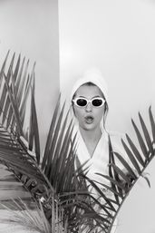Monochrome image of a woman in a bathrobe with a towel turban and retro white sunglasses peeking through large tropical palm leaves, playful surprised expression in an indoor spa-style setting.