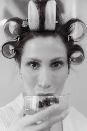 Playful black-and-white close-up of a woman with hair rollers and clips, sipping a drink while getting ready in a salon or bridal prep setting.