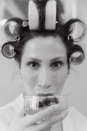 Playful black-and-white close-up of a woman with hair rollers and clips, sipping a drink while getting ready in a salon or bridal prep setting.