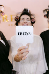 Smiling client in a beauty salon robe with hair rollers and under-eye patches holding a white handheld mirror with a logo, close-up on a manicured hand — playful salon pampering scene.