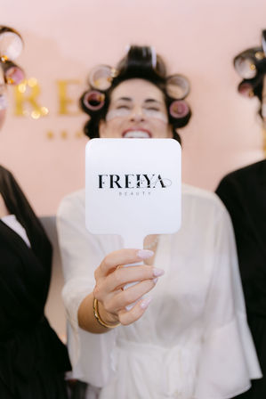Smiling client in a beauty salon robe with hair rollers and under-eye patches holding a white handheld mirror with a logo, close-up on a manicured hand — playful salon pampering scene.