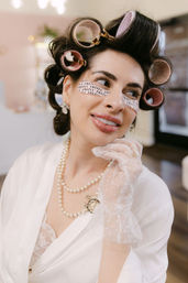 Woman in a bridal robe with pink hair rollers and under-eye patches, wearing a pearl necklace and delicate lace glove while smiling during salon bridal prep
