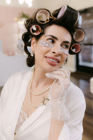 Woman in a bridal robe with pink hair rollers and under-eye patches, wearing a pearl necklace and delicate lace glove while smiling during salon bridal prep