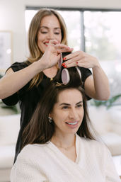 Professional hairstylist placing large velcro rollers in a smiling woman's dark hair during a salon styling appointment for a voluminous blowout.