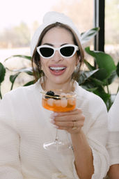 Woman in a white robe and towel wrap wearing white cat-eye sunglasses, smiling and holding an orange cocktail garnished with blackberries in a sunlit, plant-filled indoor lounge.